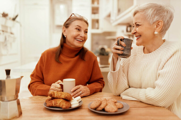 Civitas Senior Living | Portrait of stylish senior women in cozy sweaters having breakfast together at cafe, sitting at wooden table, sharing news, drinking coffee with pastries