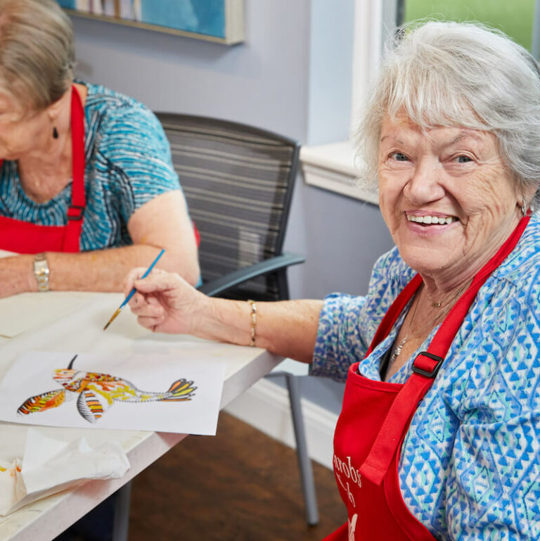 The Grand | Senior woman smiling and painting at the art table