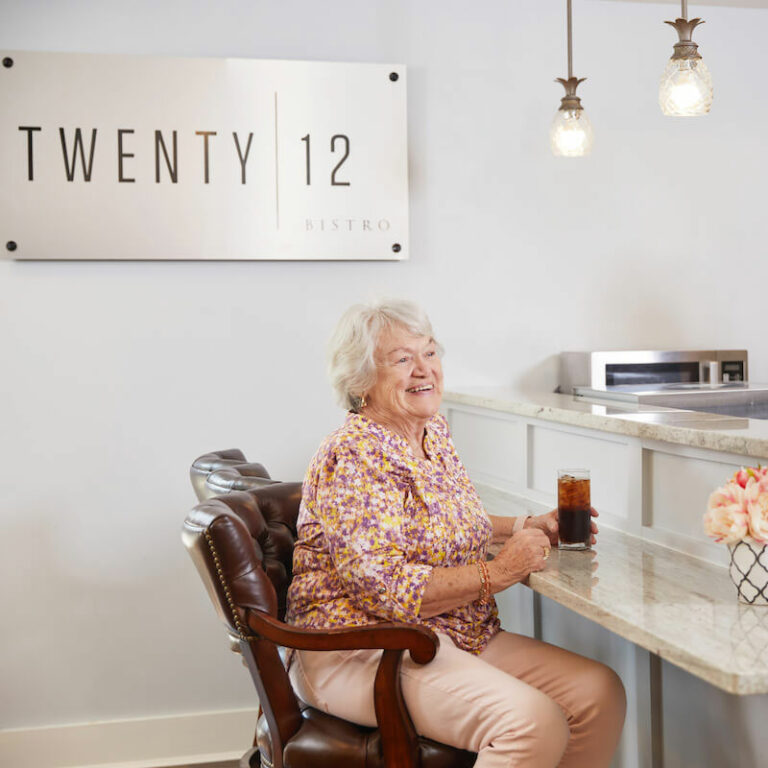 The Grand | Senior woman sitting near the reception desk talking with a caregiver and having a soda