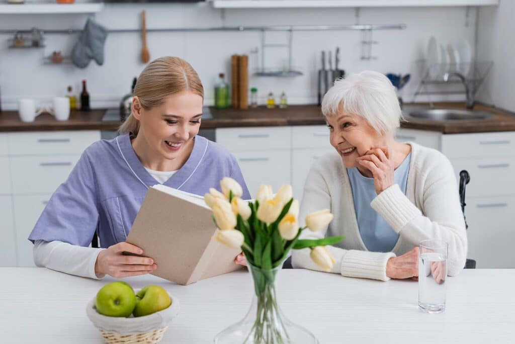 The Ridglea Senior Living | Senior woman sitting at the table with her caregiver reading a book